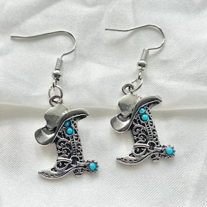 Pode incluir: Um par de brincos pendentes em tom prateado com um pingente de bota de cowboy com detalhes intrincados em filigrana e pequenos detalhes redondos na cor turquesa brilhante. Os brincos est&atilde;o presos a ganchos de metal prateado padr&atilde;o.