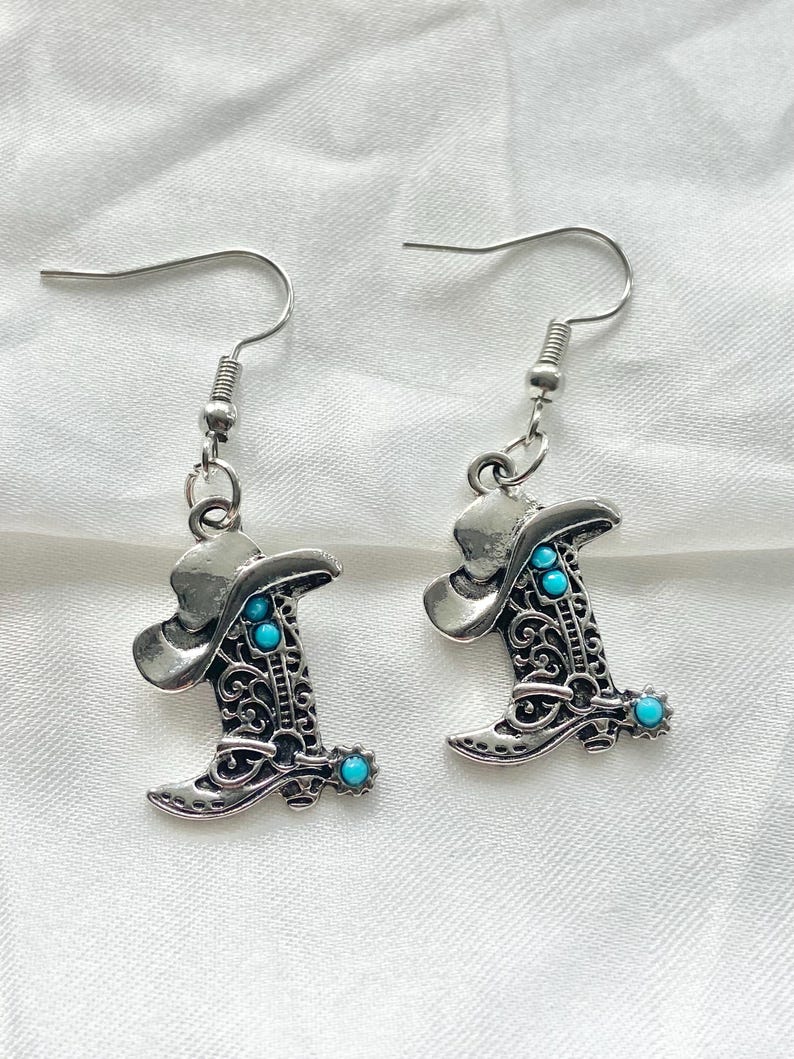 Pode incluir: Um par de brincos pendentes em tom prateado com um pingente de bota de cowboy com detalhes intrincados em filigrana e pequenos detalhes redondos na cor turquesa brilhante. Os brincos est&atilde;o presos a ganchos de metal prateado padr&atilde;o.