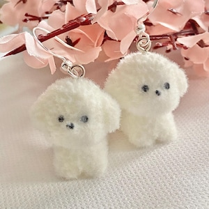 Könnte beinhalten: Ein Paar weiße, flauschige ohrringe in Hundeform mit schwarzen Perlenaugen und silberfarbenen Haken. Die Ohrringe sind vor dem Hintergrund von rosa Kirschblütenzweigen platziert.