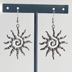 Pode incluir: Um par de brincos prateados em forma de sol. Cada brinco apresenta um centro em espiral e pontas radiantes, suspensos por um gancho simples. Os brincos são exibidos em um suporte de brinco cinza escuro sobre um fundo branco.