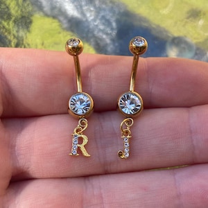 Pode incluir: Dois piercings dourados para o umbigo. Cada um tem uma gema redonda e transparente no topo e uma gema maior e transparente no centro. Um tem a letra "R" pendurada e o outro a letra "J", ambas incrustadas com pequenas gemas transparentes.
