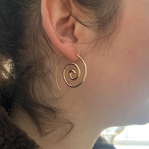 Puede incluir: Pendientes de espiral dorados. Estos pendientes presentan un diseño de espiral continuo, creando un aspecto minimalista y elegante. Los pendientes son de material dorado brillante y se muestran en una oreja.