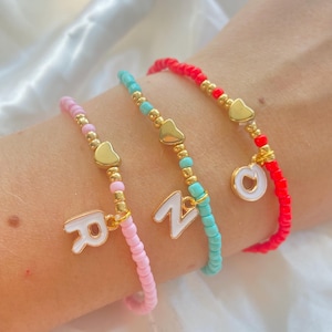 Op de afbeelding: Drie kralenarmbanden in roze, turquoise en rood. Elke armband heeft gouden hartbedels en een witte letterbedel. De armbanden zitten om een pols.