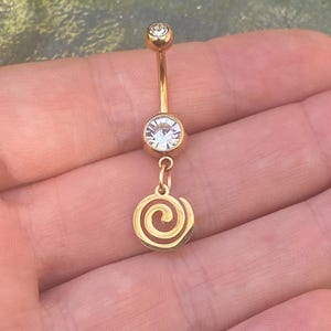 Könnte beinhalten: Ein goldfarbener Bauchnabelring mit einem klaren, runden Edelstein oben und einem größeren Edelstein in der Mitte. Ein goldener Spiralanhänger hängt am mittleren Edelstein. Der Schmuck wird in einer Hand gehalten.