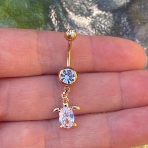 May include: A gold-toned navel ring featuring a curved barbell with a clear round crystal at the top and a larger bezel-set crystal in the centre. A dangling charm shaped like a sea turtle with a clear oval crystal shell hangs from the centre.