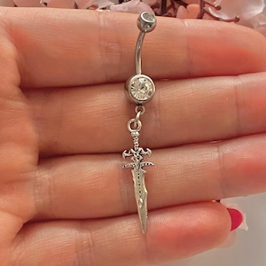 May include: A silver belly button ring with a curved barbell design. The top and bottom balls are clear, sparkling gems. A small, detailed sword charm hangs from the bottom gem, adding a unique touch to this body jewelry.