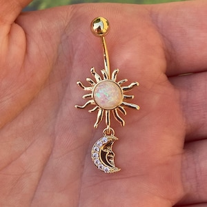 May include: Gold-colored belly button ring featuring a sunburst design with a central opal-like stone. A crescent moon charm, embellished with small clear stones, dangles below. The ring has a curved barbell and a small ball at the top.