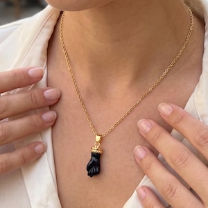 Può includere: Una collana a catena dorata con un ciondolo a forma di pugno nero. Il ciondolo presenta un cappuccio color oro nella parte superiore dove si collega alla catena. La collana poggia sul collo, evidenziando il contrasto tra l'oro e il nero.