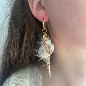 Puede incluir: Pendiente de aro dorado con un colgante de concha natural. La concha es blanca y marrón con detalles dorados y una textura espinosa. Una joya única inspirada en la naturaleza.