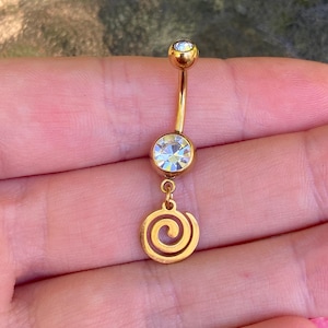 May include: A gold-toned curved barbell belly button ring featuring a small clear round gemstone at the top, a larger clear round gemstone in the center, and a dangling gold-toned spiral charm at the bottom of the jewelry piece.