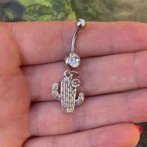 Op de afbeelding: Een zilveren navelpiercing met een gebogen staaf en twee heldere, ronde edelstenen. Een cactusbedel met een kleine bloem hangt aan de onderste edelsteen.
