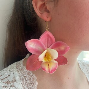 May include: Pink and white orchid flower earrings with gold-colored hooks. The earrings feature a detailed design with a yellow center and pink-tipped petals. The earrings are a floral accessory.