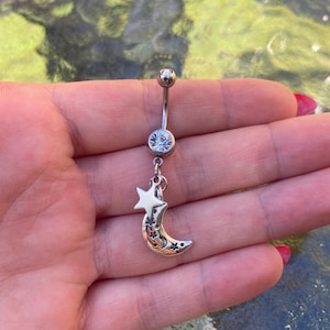 May include: A silver belly button ring with a clear gem and a dangling charm. The charm features a crescent moon with star cutouts and a star. The jewellery is held in a hand.