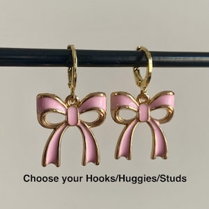 Pode incluir: Um par de brincos dourados com pingentes de laço em esmalte rosa suspensos por pequenas argolas articuladas. O texto abaixo dos brincos diz: Choose your Hooks/Huggies/Studs. Estes brincos têm um acabamento metálico polido e cor rosa.