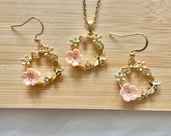 May include: A gold-coloured jewellery set comprising a necklace and earrings. Each piece features a circular design with pink flowers, small bees, and clear crystals. The set is displayed on a wooden surface.