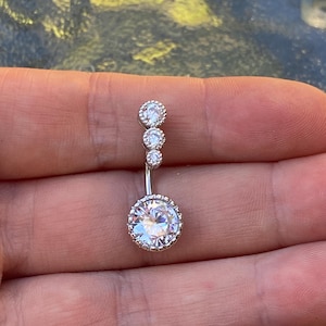 May include: A silver belly button ring featuring a large, round, clear gemstone at the base and three smaller gemstones above. The gemstones are set within a decorative, beaded border.