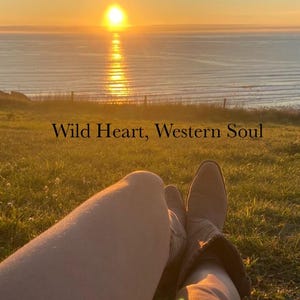 Pode incluir: Um par de botas de couro castanho-claro em estilo western cal&ccedil;adas nas pernas, posicionadas contra um p&ocirc;r do sol sobre o oceano. O texto "Wild Heart, Western Soul" est&aacute; centralizado no meio da imagem, sobrepondo a paisagem relvada.