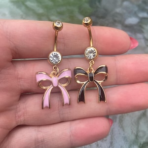Peut inclure: Deux anneaux de nombril dorés avec une tige incurvée, ornés d'un cristal rond transparent en haut et au centre. Chaque bijou comporte une breloque en forme de nœud, l'une finie en émail rose clair et l'autre en émail noir profond.