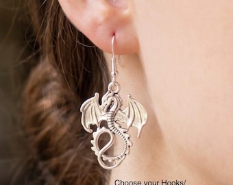 Peut inclure: Une breloque de boucle d'oreille en forme de dragon argenté suspendue à un crochet en métal. Le dragon est représenté avec les ailes déployées et une queue enroulée. Le texte "Choose your Hooks/Huggies/Studs" est imprimé en noir en bas à droite.