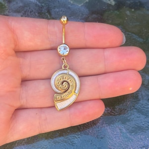 May include: A gold-toned navel ring featuring a curved barbell with a clear round crystal at the top and bottom. A dangling charm shaped like a spiral shell with white enamel inlay and intricate gold-toned detailing hangs from the bottom crystal.
