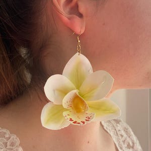 May include: A pair of large, flower-shaped earrings. The earrings feature a white and yellow orchid design with red speckles. The earrings are suspended from gold-coloured hooks. The earrings are a statement piece.