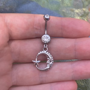 Puede incluir: Un piercing para el ombligo de barra curva en tono plateado con una pequeña piedra redonda transparente en la parte superior y una piedra redonda transparente más grande en el centro. Un dije de media luna con cara y una estrella cuelga abajo.