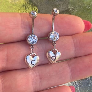 May include: Two silver-toned curved barbell belly button rings, each featuring a clear round faceted stone at the top and bottom, with a dangling heart-shaped charm. One heart is engraved with the letter C and the other with the letter Q.
