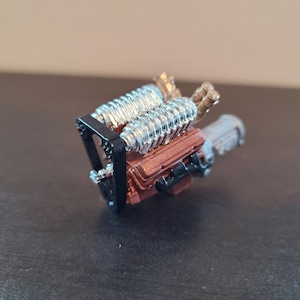 Puede incluir: Un modelo detallado de un motor V8 con partes pintadas en plata y oro. El motor está montado en un soporte de metal negro.