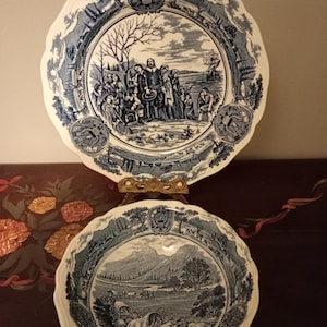 Puede incluir: Dos platos antiguos de cerámica azul y blanca. El plato superior presenta una escena detallada de personas cerca de la costa, con un barco al fondo. El plato inferior representa un paisaje con carretas cubiertas y ganado.