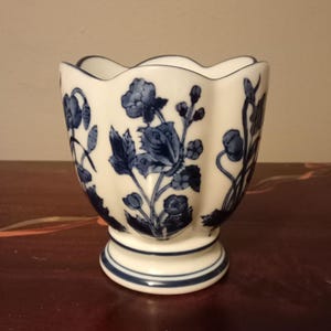 Pode incluir: Um vaso de cerâmica branca com borda ondulada e base de pedestal. O vaso é decorado com um padrão floral azul. A base e a borda são acentuadas com acabamentos azuis. O vaso é provavelmente para fins decorativos.