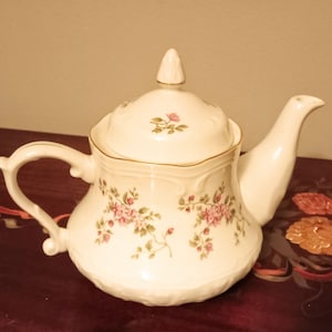 Puede incluir: Una tetera de cerámica color crema con un delicado patrón floral de pequeñas flores rosas y hojas verdes. La tetera tiene una tapa a juego con un remate puntiagudo, un asa curva y un pico, todo acentuado con un fino borde dorado en los bordes.