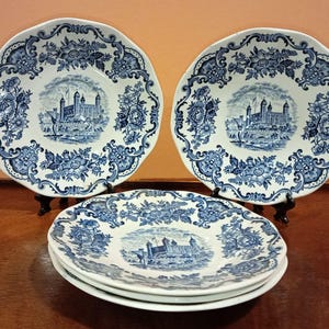 Può includere: Un set di piatti in ceramica bianca con un design floreale e architettonico blu. I piatti presentano un'immagine centrale di un castello e sono disposti su una superficie scura. I piatti sono in stile classico.