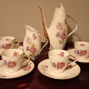 May include: A white porcelain tea set with floral designs. The set includes a tall coffee pot, a creamer, and four teacups with saucers. The cups, saucers, and creamer have gold trim. The floral design features red, purple, and yellow flowers.