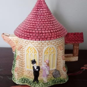 May include: A ceramic church-shaped teapot with a pink tiled roof and a gold cross. The teapot features a brick facade, arched windows, and figures of a bride and groom. A decorative item.