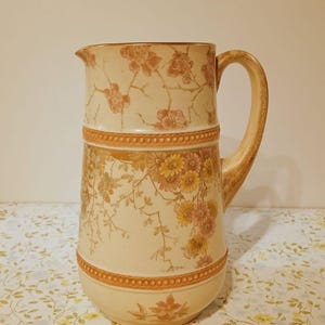 May include: A ceramic pitcher featuring a cream-colored base with a floral pattern in shades of orange, yellow, and brown. The vessel includes a curved handle, a pouring spout, and two horizontal bands with a raised, beaded texture around the body.