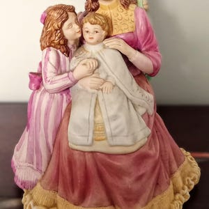 Könnte beinhalten: Porzellanfigur, die eine Frau und zwei Kinder darstellt. Die Frau trägt ein rosa Kleid mit goldenem Besatz. Die Kinder tragen weiße und rosa Outfits. Die Figur hat ein detailliertes Design und eine Vintage-Ästhetik.