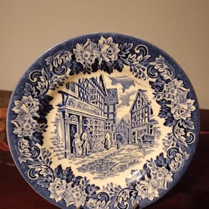 May include: A decorative plate with a blue and white floral border. The center features a detailed illustration of a street scene with buildings and figures. The plate has a glossy finish and is displayed on a gold-colored stand.
