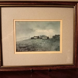 Könnte beinhalten: Gerahmtes Aquarell-Landschaftsbild, das eine ländliche Szene mit Gebäuden auf einem Hügel unter einem bewölkten Himmel zeigt. Das Kunstwerk ist in einem dunkelbraunen Holzrahmen mit einem hellen Passepartout gerahmt.