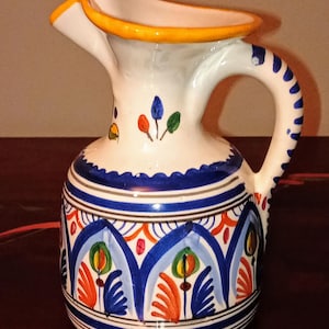 Può includere: Brocca in ceramica bianca con manico e beccuccio curvi. La brocca è decorata con motivi floreali blu, arancioni e verdi e un manico a righe blu e bianche. Il bordo è rifinito con giallo.