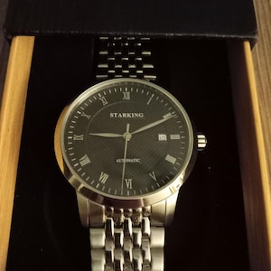 May include: A silver-toned Starking automatic wristwatch with a black face and Roman numeral hour markers. The watch has a date display and a stainless steel band. The watch is presented in a black box with a wooden frame.