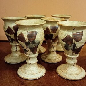 May include: A collection of six ceramic goblets with a speckled beige finish. Each goblet features a wide base, a slender stem, and a flared rim. Dark brown leaf-like patterns adorn the exterior of each piece, adding a touch of rustic charm.
