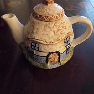 May include: A cream-coloured ceramic teapot, designed to resemble a cottage with a thatched roof. It has small windows, a door, and a handle. The teapot is decorated with brown and blue details.