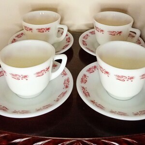 May include: Four white teacups and saucers with a red floral design. The cups have handles and sit on matching saucers. The set is arranged on a dark wooden surface, creating a classic and elegant aesthetic.