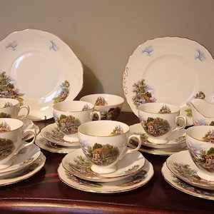 May include: A vintage tea set featuring white porcelain cups, saucers, bowls, and plates. Each piece is decorated with a scenic landscape design and gold trim. The set includes teacups, saucers, bowls, serving plates, and a creamer.