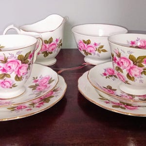 May include: A traditional tea set comprising two teacups with saucers, a creamer jug, and a sugar bowl. The white porcelain is adorned with pink rose motifs and gold edging. A charming set for afternoon tea.
