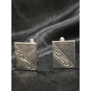 May include: A pair of silver-toned cufflinks. Each cufflink is rectangular with a diagonal etched design and a small, rounded post. The background is a dark, textured fabric.