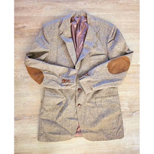 May include: A tan and brown herringbone blazer featuring brown suede elbow patches, a notched lapel, two front buttons, and three smaller buttons on each sleeve cuff. The jacket is lined with a smooth, solid brown fabric and includes two front pockets.