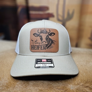 May include: Tan and white trucker hat with a leather patch featuring a cow graphic and the words "Horny Heifer." The hat has a curved brim and an adjustable strap. The Richardson 112 hat is a classic trucker style.