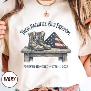 May include: Ivory-colored t-shirt featuring a graphic of combat boots, a folded American flag, and the text "Their Sacrifice. Our Freedom. Forever Honored - 1776 to 2026." The shirt also has the word "IVORY" printed on it.