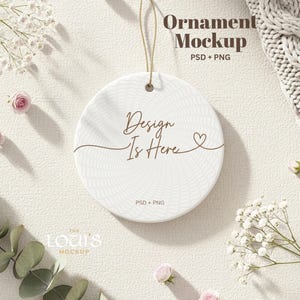 May include: A round white ornament with the text "Design Is Here" in brown script, with a heart symbol. The ornament is hanging by a gold string. The background includes flowers and the text "Ornament Mockup".